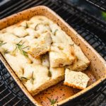 Focaccia mit Rosmarin in einer rechteckigen, gesprenkelten Form auf einem Grillrost, einige Stücke herausgeschnitten.