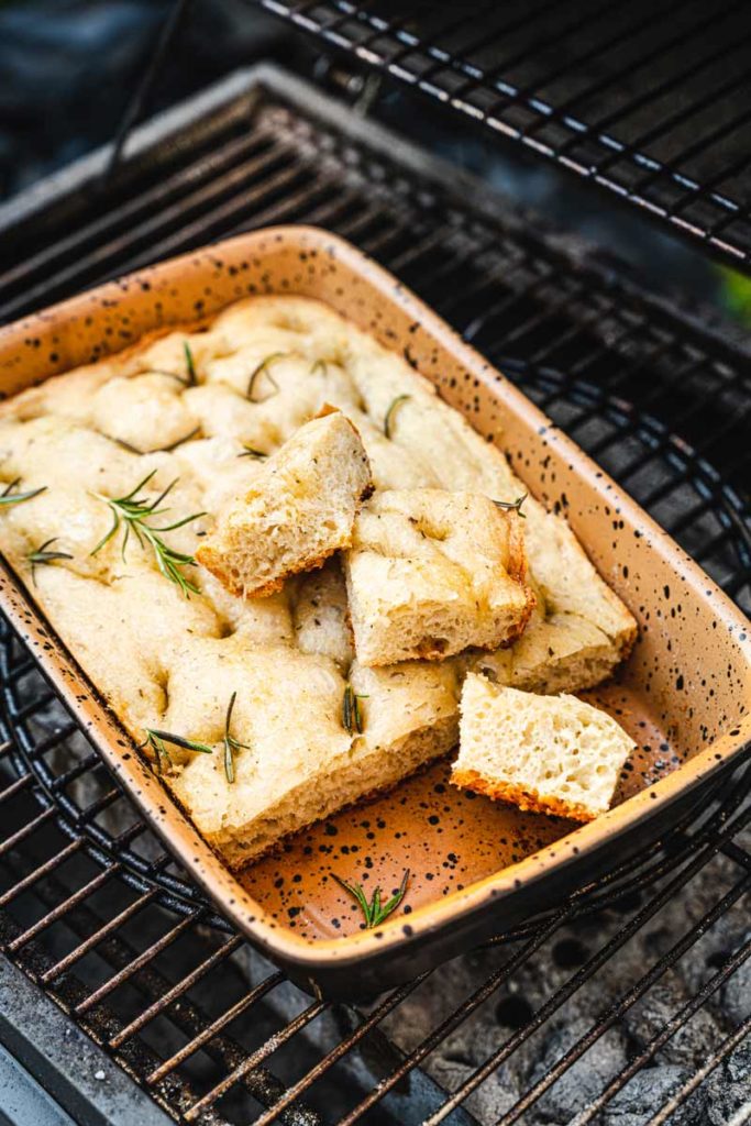 Focaccia mit Rosmarin in einer rechteckigen, gesprenkelten Form auf einem Grillrost, einige Stücke herausgeschnitten.
