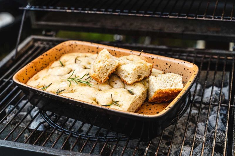 Focaccia mit Rosmarin in einer Auflaufform auf einem Grill, mehrere Stücke sind herausgeschnitten.