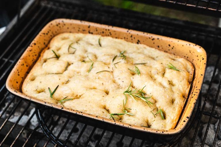 Focaccia in einer orangefarbenen Keramikform wird auf einem Grill gebacken, garniert mit frischem Rosmarin.