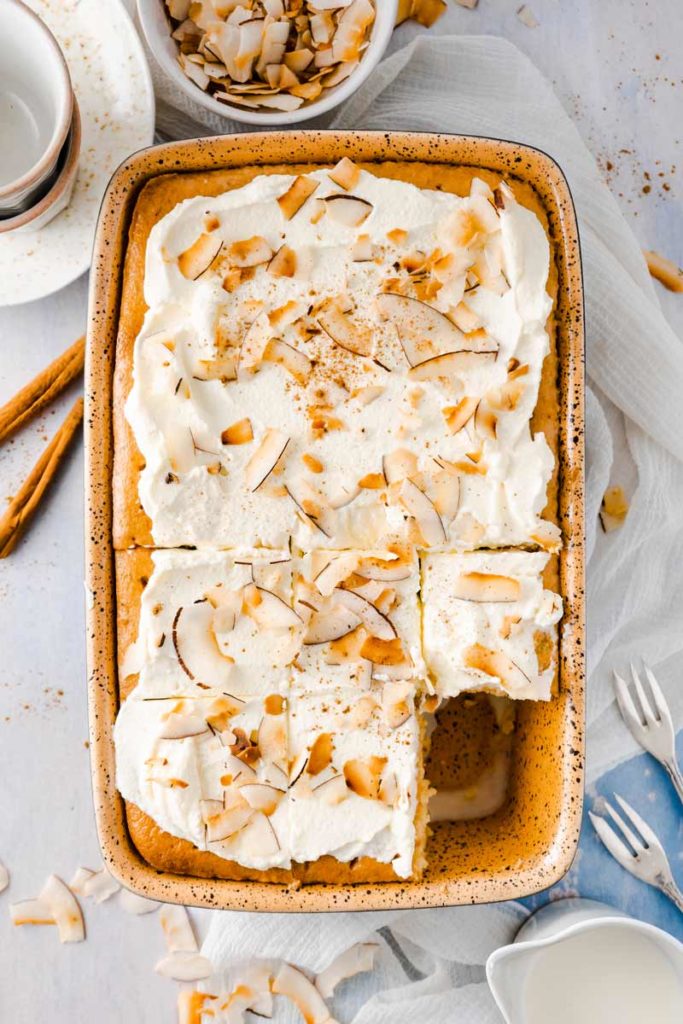 Blech mit Kuchen, bedeckt mit Sahne und Kokoschips, in Quadrate geschnitten. Daneben Zimtstangen und Besteck.