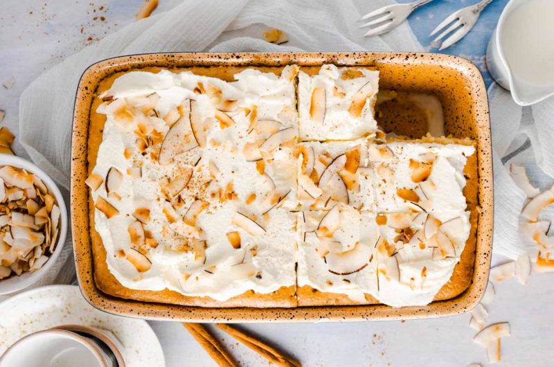 Kuchen in Backform, bedeckt mit Sahne und Kokosraspeln, daneben Gabeln, Milch und Kokoschips.