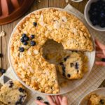 Blaubeer-Streuselkuchen auf Teller, mit Mandelscheiben garniert; ein Stück fehlt, Hände greifen nach einem weiteren Stück.