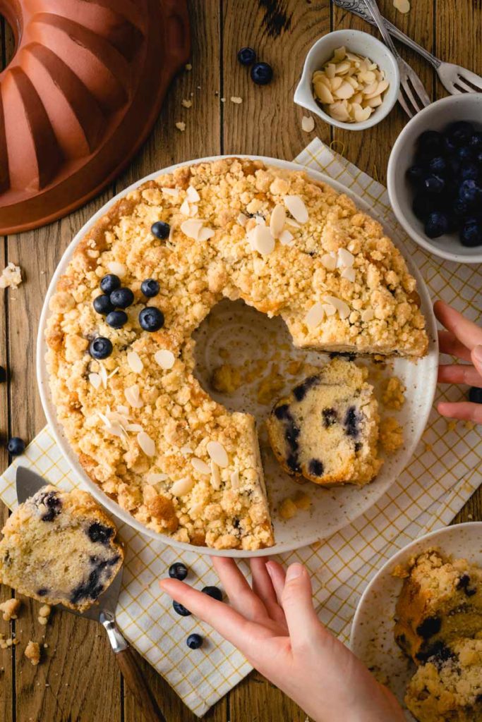 Blaubeer-Streuselkuchen auf Teller, mit Mandelscheiben garniert; ein Stück fehlt, Hände greifen nach einem weiteren Stück.