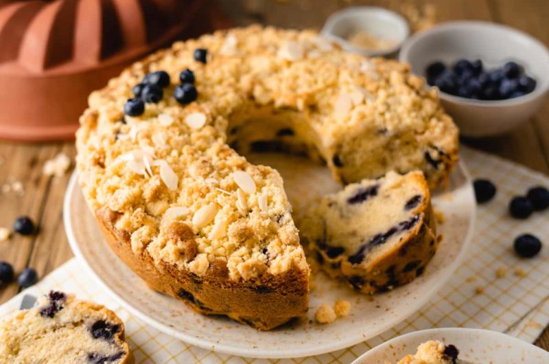 Kuchen mit Heidelbeeren und Streuseln auf Teller. Eine Scheibe fehlt. Im Hintergrund sind Schalen mit Beeren.