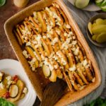 Auflaufform mit geschichtetem Kartoffel-Zucchini-Gericht, garniert mit Feta. Daneben frische Tomaten und Basilikum.