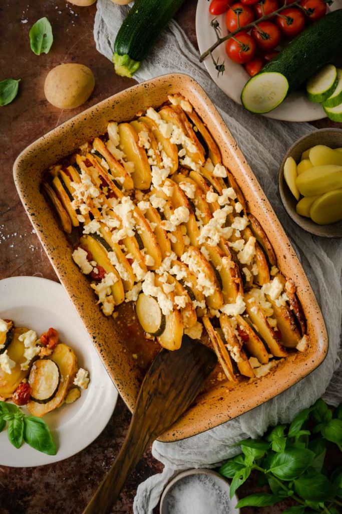Auflaufform mit geschichtetem Kartoffel-Zucchini-Gericht, garniert mit Feta. Daneben frische Tomaten und Basilikum.