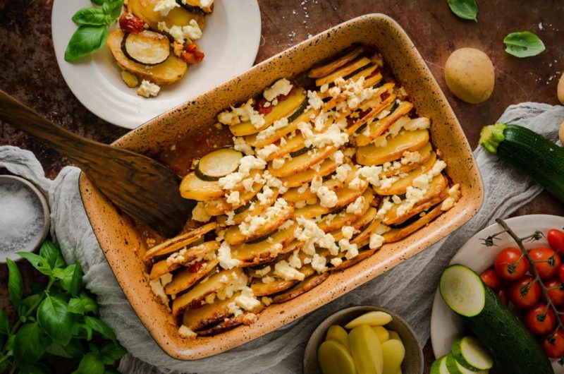 Kartoffel-Zucchini-Auflauf mit Feta in einer Auflaufform, umgeben von frischen Zutaten wie Tomaten und Basilikum.