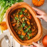 Ein Auflauf mit Kürbis und Fleisch in einem Keramiktopf, dekoriert mit Petersilie, umgeben von Kürbissen und Kräutern.