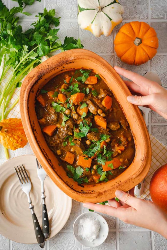 Ein Auflauf mit Kürbis und Fleisch in einem Keramiktopf, dekoriert mit Petersilie, umgeben von Kürbissen und Kräutern.