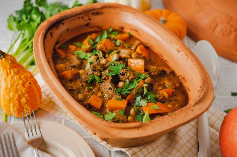 Herbstlicher Eintopf im Tontopf mit Kürbis, Kichererbsen und Kräutern, umgeben von herbstlicher Dekoration.