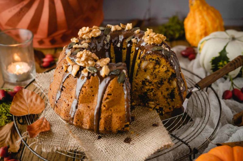 Herbstlicher Gugelhupf mit Schokoglasur, Nüssen und Kürbiskernen auf einem Gitter, umgeben von Kerzen und Herbstdeko.
