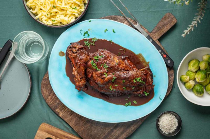 Bratenteilstück auf blauem Teller, mit Soße, umgeben von Schüssel Spätzle und Rosenkohl, auf grünem Tischtuch.