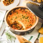 Auflaufform mit gebackenem Shepherd's Pie und Petersilie, daneben Kartoffeln und ein Stück auf einem Teller.