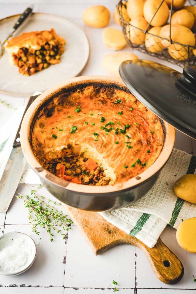 Auflaufform mit gebackenem Shepherd's Pie und Petersilie, daneben Kartoffeln und ein Stück auf einem Teller.
