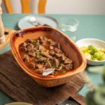 Eintopf im Keramiktopf mit Spätzle und Rosenkohl auf einem Holztisch, gedeckt mit Teller und Glas.