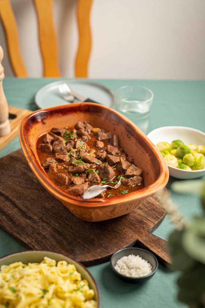 Eintopf im Keramiktopf mit Spätzle und Rosenkohl auf einem Holztisch, gedeckt mit Teller und Glas.
