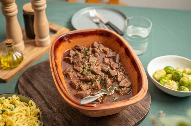 Eintopf im Tontopf, serviert mit Schüssel Spätzle und Rosenkohl, auf grünem Tisch mit Wasser und Gewürzen.