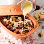 Keramikschale mit Brot-Pudding und Beeren, übergossen mit Vanillesoße. Im Hintergrund Brotwürfel und Tischdeko.
