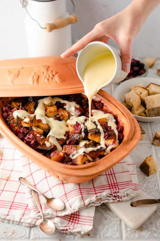 Keramikschale mit Brot-Pudding und Beeren, übergossen mit Vanillesoße. Im Hintergrund Brotwürfel und Tischdeko.