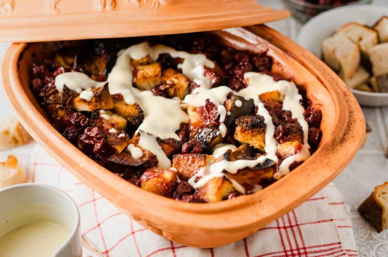 Auflaufform mit Brotwürfeln, Kirschen und Vanillesoße auf einem rot-weißen Tuch, im Vordergrund ein Becher.