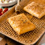 Zwei Sesamgebäckstücke auf einem Backblech mit Honigbesen, daneben Tomaten und ein Salat im Hintergrund.