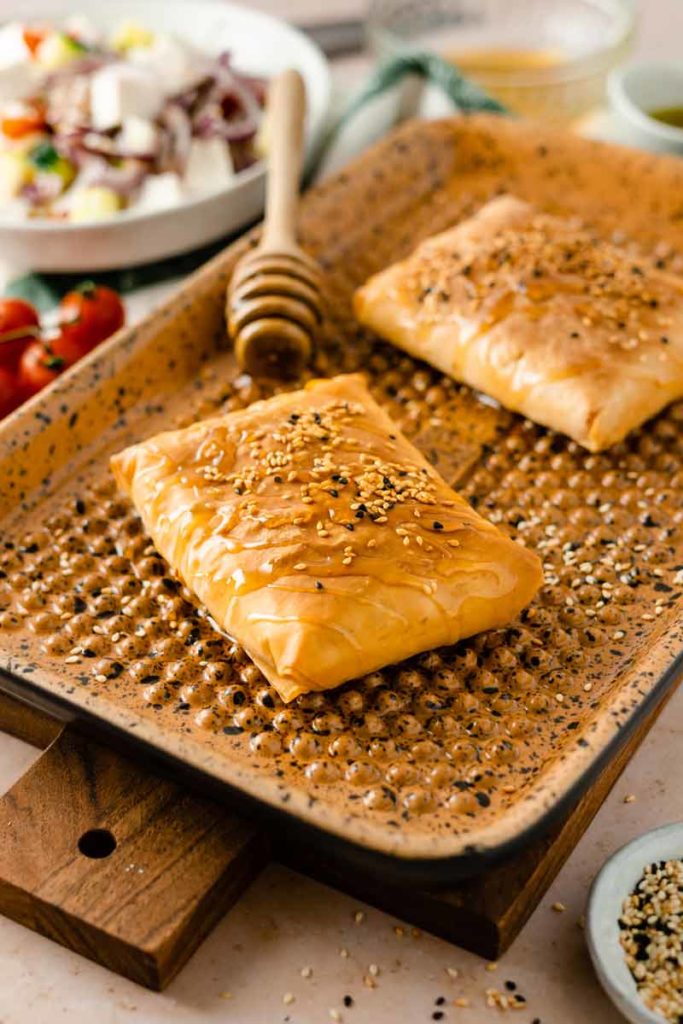 Zwei Sesamgebäckstücke auf einem Backblech mit Honigbesen, daneben Tomaten und ein Salat im Hintergrund.