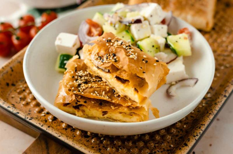 Blätterteigkuchen mit Sesam und griechischem Salat in einer weißen Schüssel auf einem Tablett.
