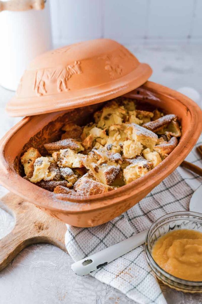 Brauner Tontopf mit Kaiserschmarrn, bestäubt mit Puderzucker; daneben eine Schale Apfelmus auf einem Holzbrett.