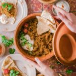 Hummusschale mit Fladenbrot, Joghurt und Koriander auf Tisch, rote Linsen in Holzschale, Teller mit Hummus-Garnitur und Messer.