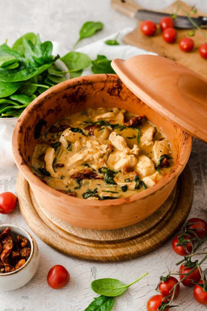 Tontopf mit cremigem Hähnchengericht, Spinat, Tomaten und Gewürzen, umgeben von frischen Zutaten auf Holzbrett.
