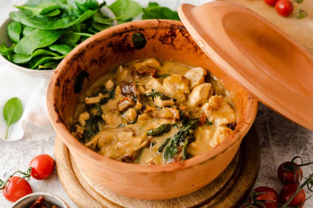 Keramiktopf mit cremigem Hühnchen, Spinat und sonnengetrockneten Tomaten. Frische Tomaten und Spinatblätter daneben.
