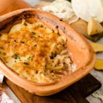 Auflauf in brauner Form mit geschmolzenem Käse, daneben geschnittenes Gemüse und ein Messer auf einem Holzbrett.