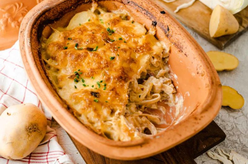 Überbackener Kartoffelauflauf in einer Keramikform, bestreut mit frischen Kräutern, daneben Kartoffeln auf einem Handtuch.