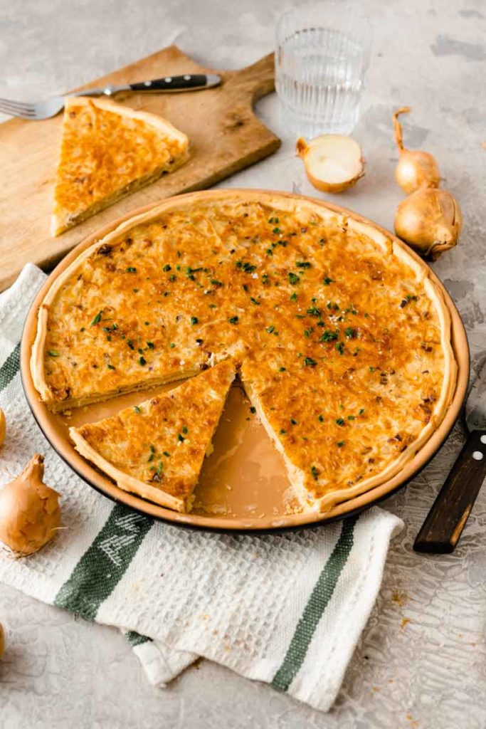 Zwiebelkuchen auf einem Teller mit einem angeschnittenen Stück, daneben ein Holzbrett mit einem weiteren Stück.