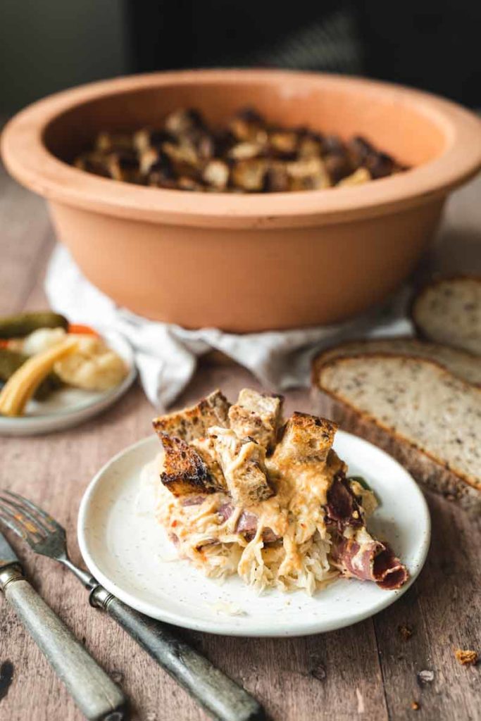 Ein Teller mit Sauerkraut, Fleisch und Brotstücken vor einem großen Tontopf und eingelegtem Gemüse.