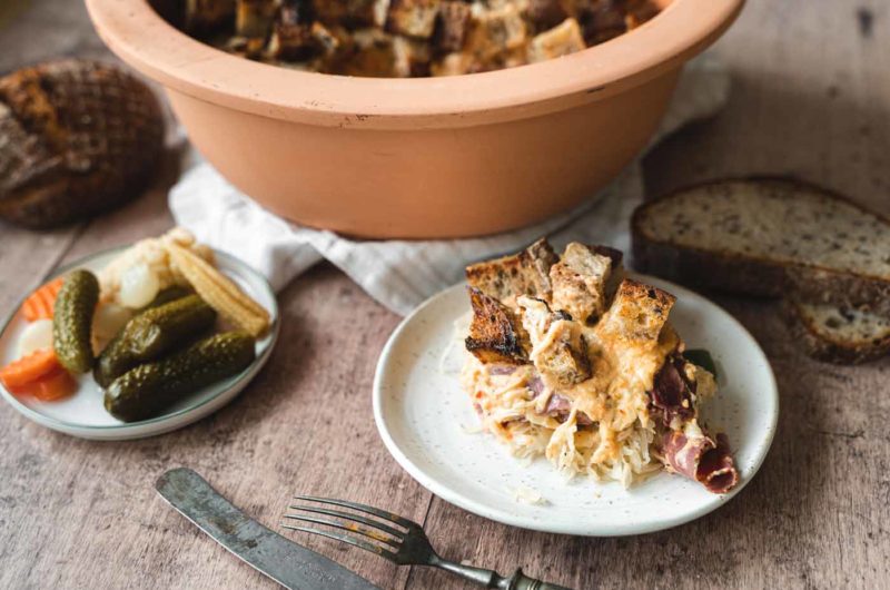 Teller mit herzhaftem Auflauf aus Sauerkraut, Brot und Fleisch. Daneben Teller mit Mixed Pickles und Brotscheiben im Hintergrund.
