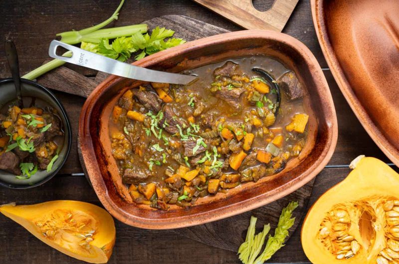 Eintopf mit Kürbis und Rindfleisch in einem Keramiktopf. Frische Kräuter und geschnittene Kürbisstücke daneben.