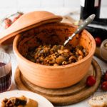 Terrakotta-Schüssel mit Bohneneintopf, daneben ein Glas Rotwein und Ciabatta-Scheiben auf einem Teller.
