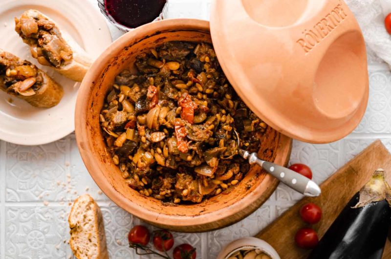 Ton-Schmortopf mit mediterranem Gemüseeintopf, Brot, Tomaten und einem Glas Rotwein auf weißem Tisch.