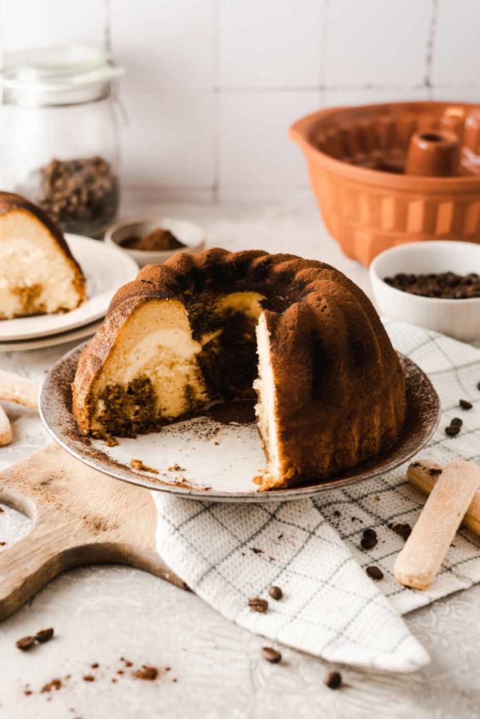 Anschnitt eines Gugelhupfs auf Teller, im Hintergrund Löffelbiskuits und Kaffeebohnen.