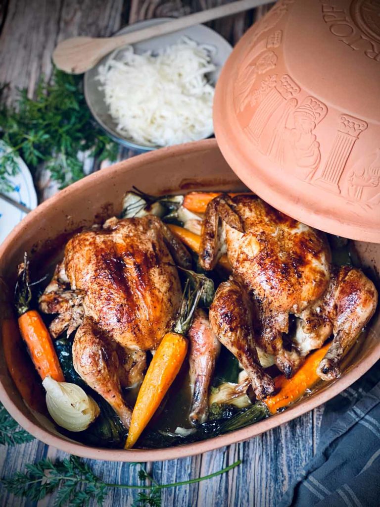 Zwei gebratene Hähnchen mit Karotten und Zwiebeln in einem Tontopf, daneben eine Schüssel mit geriebenem Käse.