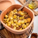 Tonform mit Pasta, Hähnchen und getrockneten Tomaten, daneben Gewürzöl und frische Tomaten.
