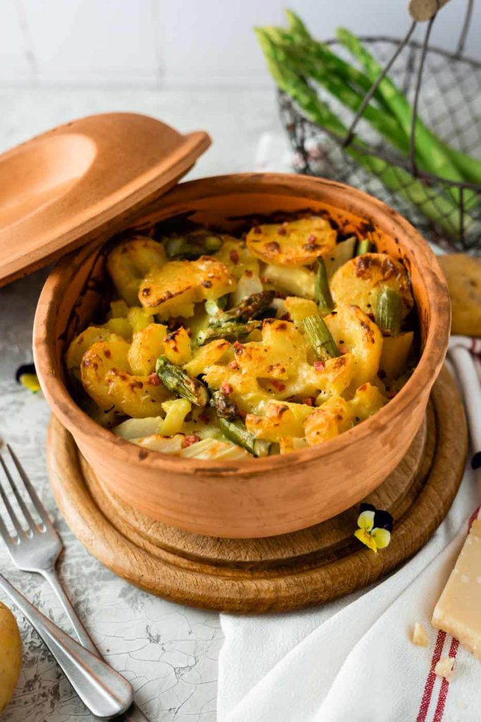 Ofen-Gemüse mit Spargel in einem runden Tontopf, daneben Besteck und eine Blume auf einem Serviettentuch.