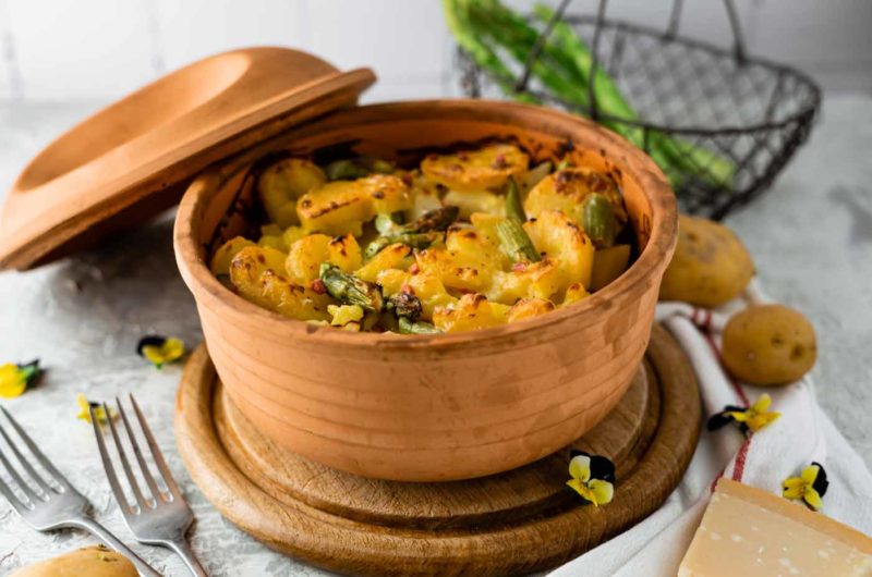 Kartoffelauflauf mit Spargel in einem Tontopf, serviert auf einem Holzteller, mit Gabeln und Parmesan auf einem Tuch.