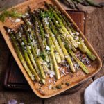 Grüner Spargel im Ofen gebacken, garniert mit geriebenem Käse und Kräutern in einer Keramikschale auf Holztisch.