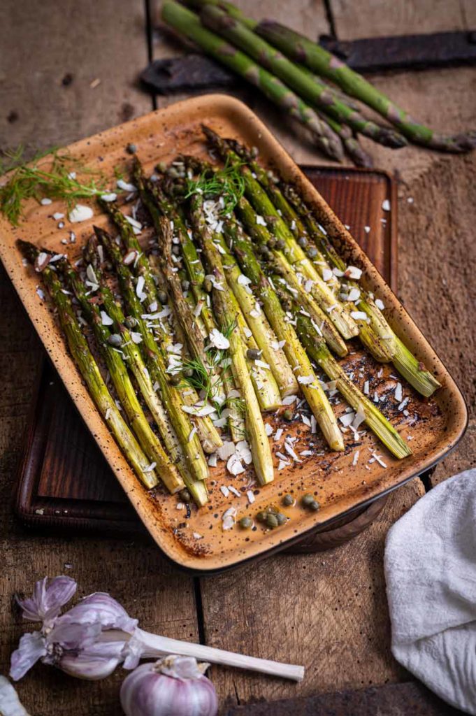 Grüner Spargel im Ofen gebacken, garniert mit geriebenem Käse und Kräutern in einer Keramikschale auf Holztisch.