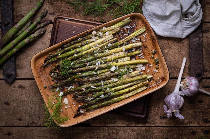 Gerösteter grüner Spargel mit Mandelscheiben und Kräutern auf einem Holzbrett, daneben Knoblauch und ein Tuch.
