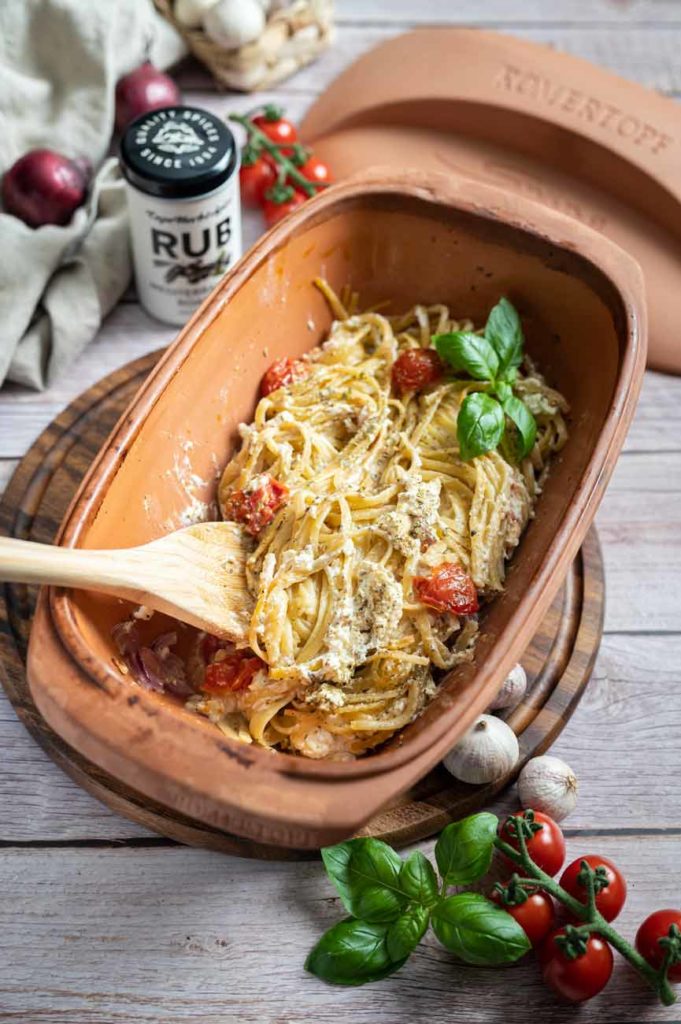 Nudelgericht mit Tomaten und Basilikum in Tonauflaufform, daneben Gewürz, Kirschtomaten und Knoblauch auf Holzbrett.