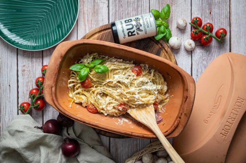 Gebackene Spaghetti mit Tomaten und Kräutern in einer Tonform, umgeben von frischen Zutaten und einem Gewürzglas.